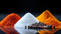 Vibrant piles of colorful spices and seasonings including red chili powder, white salt, turmeric powder and black peppercorns arranged on dark reflective surface.