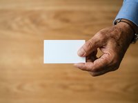 Male hand with darker skin tone wearing light blue sleeve holding blank white card against wooden background, suitable for business card mockups, corporate presentations, and professional branding materials.