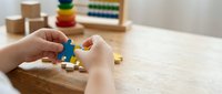 Child hands playing with colorful wooden puzzle pieces on table in bright educational playroom with learning toys and abacus in background.