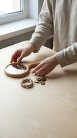 Person in linen clothing arranging coins and a wooden hand mirror on a light wooden table, with a hair tie nearby, in a calm minimal interior.