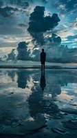 Male silhouette standing on wet beach at sunset with dramatic storm clouds and mirror reflection in water, creating surreal atmospheric seascape scene.