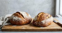 Two freshly baked artisan sourdough bread loaves with golden crusts on wooden cutting board in rustic kitchen setting for bakery and cooking concepts.