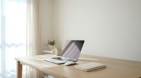 Clean minimalist workspace with open laptop on wooden desk near bright window. Modern home office setup with natural light and neutral decor for productivity and focus.