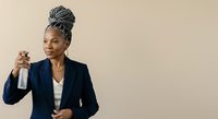 Middle-aged African American woman in professional navy blazer holding spray bottle, promoting cleaning services, household products, or sanitization solutions against neutral background.