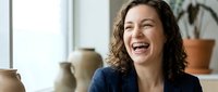 Young professional woman with curly brown hair laughing joyfully in bright modern office workspace with pottery and plants in background.