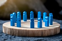 Blue wooden figures standing in circle on natural wood slice platform against dark background, representing team meeting, unity and collaboration concept.