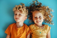Cheerful Caucasian children with curly blonde hair lying on turquoise background wearing bright orange and yellow t-shirts, expressing joy and playfulness.
