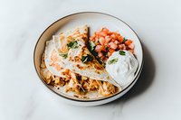 Pulled pork tacos with fresh coleslaw, diced tomatoes and sour cream served on white corn tortillas, garnished with cilantro on ceramic plate.