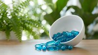 White bowl spilling blue gel capsules on wooden surface with green plants in background for health and wellness supplement concepts.