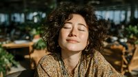 Young woman with curly hair enjoying peaceful moment with closed eyes and gentle smile in modern cafe or restaurant setting with natural lighting.