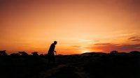 Young man in silhouette standing on rocky terrain during golden hour sunset, gazing toward horizon with contemplative posture, suitable for motivational and lifestyle content.