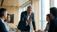 Mature male executive with gray hair leading business meeting with diverse colleagues in modern office environment with natural light from windows.