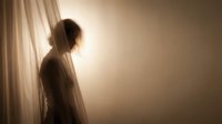 Young woman with long hair standing by window with sheer curtains, backlit by warm sunlight, creating silhouette effect. Contemplative mood for wellness, mindfulness, and lifestyle concepts.