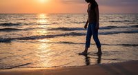 Young woman walking barefoot on sandy beach at golden sunset with ocean waves reflecting warm light creating peaceful seaside moment for relaxation and wellness.