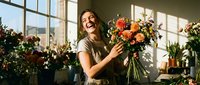 Young woman with long brown hair and fair complexion arranging fresh orange flowers in a bright studio florist workspace surrounded by potted plants and natural window light.