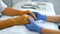 Doctor in blue gloves holding elderly patient hands during medical consultation, showing care and support in clinic.