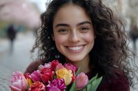Young Hispanic woman with curly dark hair smiling brightly while holding colorful spring tulip bouquet featuring pink, red and yellow blooms against blurred urban background.