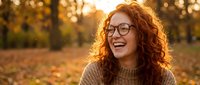 Young woman with curly red hair and glasses laughing joyfully in autumn park during golden hour sunset. Natural happiness and positive emotions outdoors.