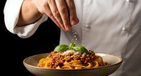 Professional chef adding fresh herbs to homemade pasta with rich tomato meat sauce in rustic ceramic bowl for gourmet Italian cuisine presentation.