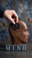 Hand inserting gear into wooden head sculpture with brain gears engraved, metal blocks spelling mind below, representing mental mechanics, cognitive development, psychology, and innovative thinking concepts.