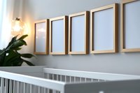 Modern nursery interior with row of empty gold frames on light gray wall above white crib, decorative plant and wall sconce creating cozy atmosphere.
