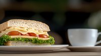 Fresh sandwich with cheese, tomato, lettuce on whole grain bread served on white plate next to coffee cup against dark background, breakfast or lunch setting.