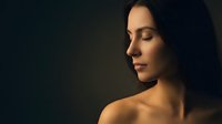 Young woman with closed eyes in dramatic lighting showing peaceful expression and bare shoulders against dark background for wellness and beauty concepts.
