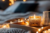 Cozy winter evening atmosphere with glowing candle in decorative glass holder on wooden tray, soft blanket and warm bokeh lights creating relaxing ambiance.