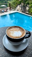 Hot cappuccino with latte art foam served in black and white ceramic cup on stone surface overlooking luxurious swimming pool surrounded by tropical greenery.