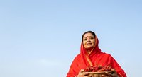 Woman wearing red traditional headscarf and clothing holding basket of fresh produce against clear blue sky background for cultural lifestyle and agriculture concepts.