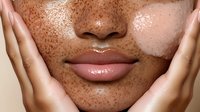 Young woman with freckles applying skincare treatment, hands gently touching face with glowing dewy skin for beauty and wellness routine.