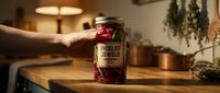 Hand holding jar of pickled red vegetables with label on wooden table in warm kitchen setting with dried herbs and soft lighting.