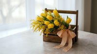 Yellow tulips and mimosa flowers in rustic wooden basket with beige ribbon bow on table near window for spring home decor and gift arrangements.