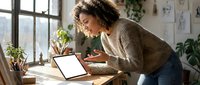 Young woman with curly hair and medium skin tone working at wooden desk with laptop, notebook and pencil holder in bright home office with plants and window light.