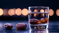 Fresh dates soaking in clear water glass with blurred bokeh lights in background, dark moody food photography setup on black surface.