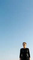 Woman in black patterned dress standing against clear blue sky, looking upward with contemplative expression. Minimal composition with ample negative space for design and text overlay.