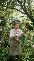 Middle-aged Caucasian female gardener in apron and work gloves tending pink roses in lush garden setting with mature trees and flowering plants overhead.