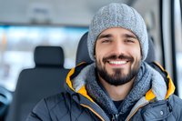 Young bearded Caucasian man wearing winter clothing smiles warmly while sitting in car. Gray knit beanie, yellow puffer jacket and genuine expression create welcoming atmosphere.
