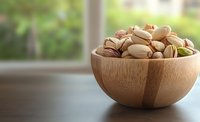 Fresh roasted pistachios in wooden bamboo bowl on dark table against blurred window background, healthy snack food photography with copy space.