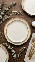 Elegant table setting with cream plates trimmed in gold, fresh eucalyptus and rosemary sprigs, linen napkin, and gold cutlery on wooden surface. Perfect for wedding, dinner party, or hospitality design projects.