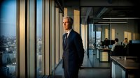 Middle-aged Caucasian male professional in dark business suit standing in modern high-rise office corridor with floor-to-ceiling windows overlooking cityscape, suitable for corporate, leadership, and business concepts.