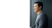 Asian male professional in gray sweater leaning against modern wall, contemplative expression, suitable for corporate, business, lifestyle, and editorial projects.