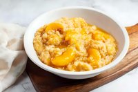 Creamy oatmeal porridge topped with fresh peach slices and honey in white ceramic bowl on wooden board against marble background, morning breakfast setup.