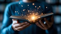 Open book emitting magical glowing light particles and sparkles in hands wearing blue sweater against dark background, creating mystical reading atmosphere.