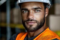 Close up portrait of confident construction worker in orange safety vest and hard hat with intense blue eyes and trimmed beard looking at camera.
