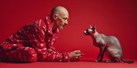Bald Caucasian man in red patterned pajamas playfully interacting with hairless sphynx cat on matching red background for lifestyle advertising.