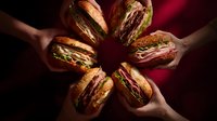 Hands holding delicious sandwiches arranged in circle against dark red background, showcasing variety of fresh ingredients in artisanal bread.