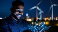 Young African American engineer holding glowing digital network tablet at wind farm during evening hours showcasing renewable energy technology and sustainable power solutions.
