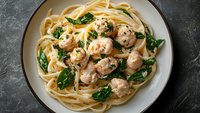 Creamy fettuccine pasta with tender chicken meatballs and fresh spinach leaves in garlic sauce served on gray ceramic plate against dark stone background.