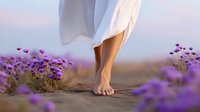 Barefoot woman in white dress walking through blooming purple wildflowers at sunset, low angle view showing feet and dress hem against soft pastel sky.